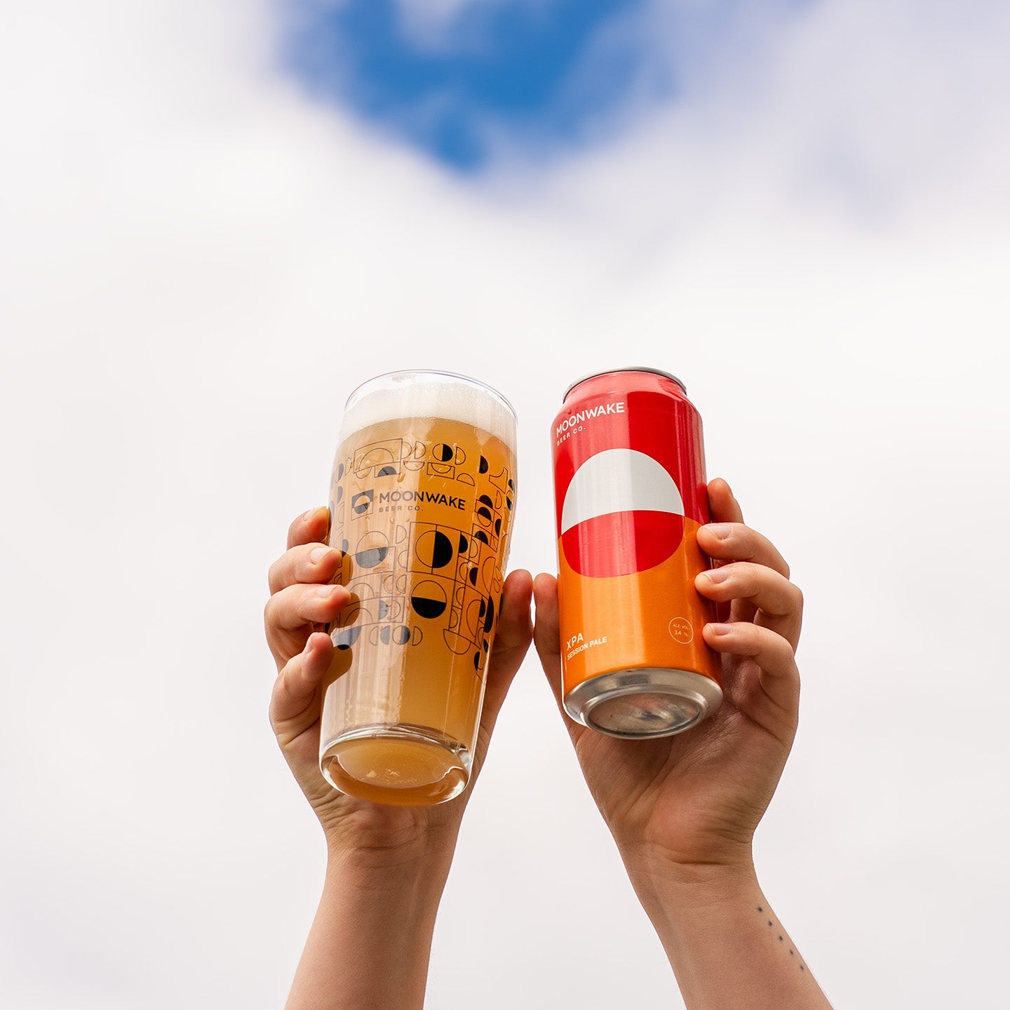 Two hands holding a frothy glass of Moonwake XPA beer and a red and orange can of XPA against a cloudy blue sky background.