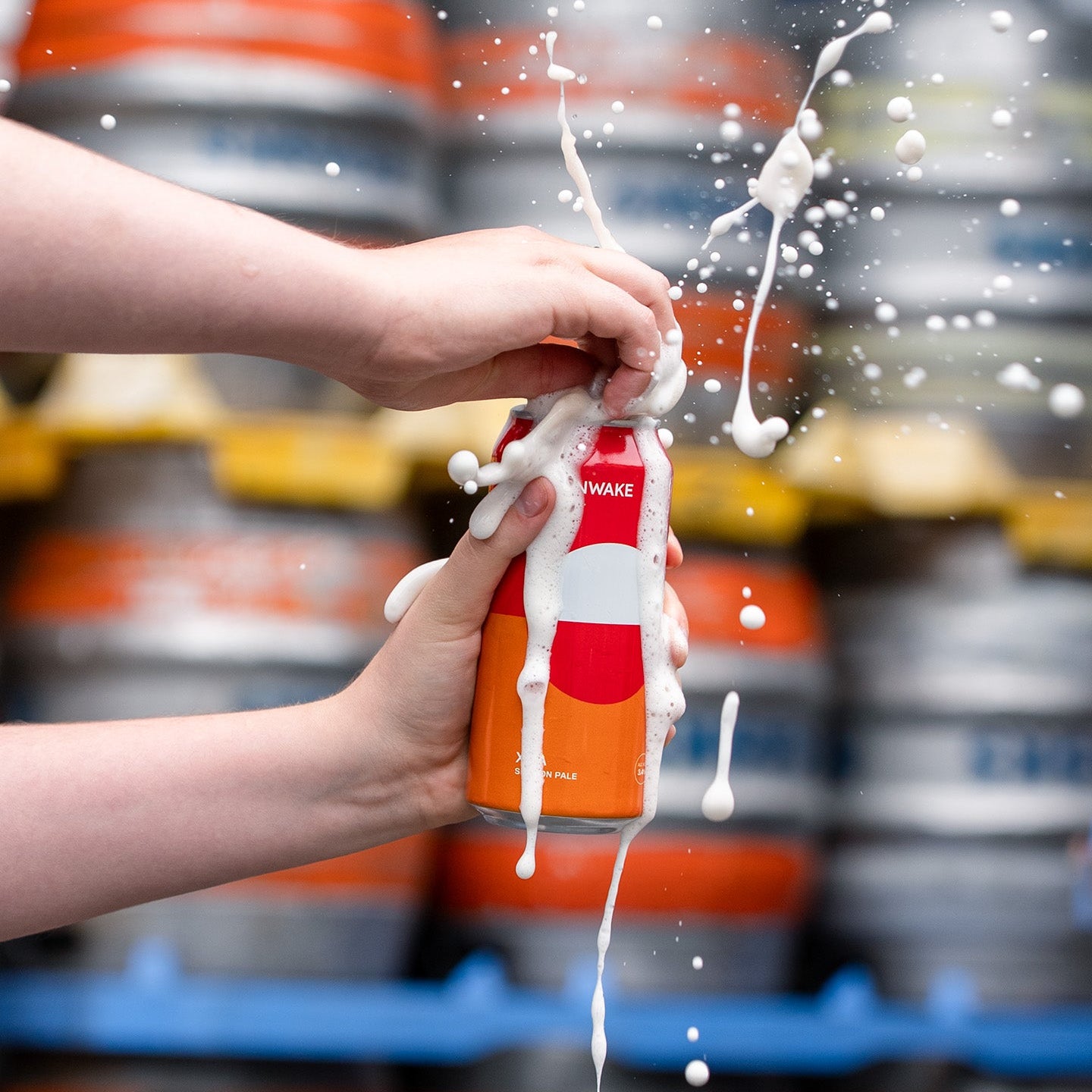 A person's hands holding an orange and white can of Moonwake XPA beer, with white foam spraying dramatically in the air against a blurred background of stacked kegs.