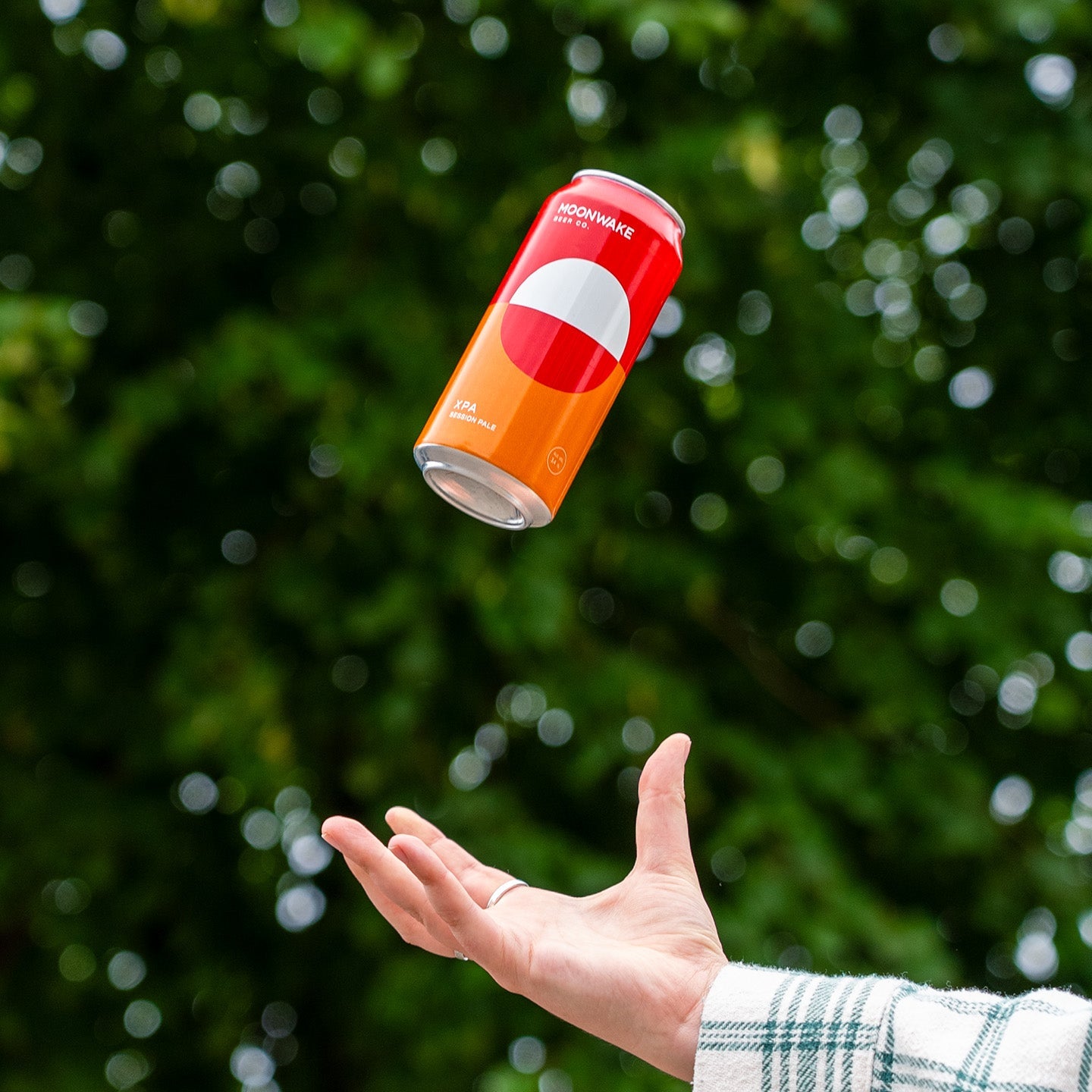 A red and orange can of Moonwake XPA floating in mid-air above an open hand, with a blurred green outdoor background.